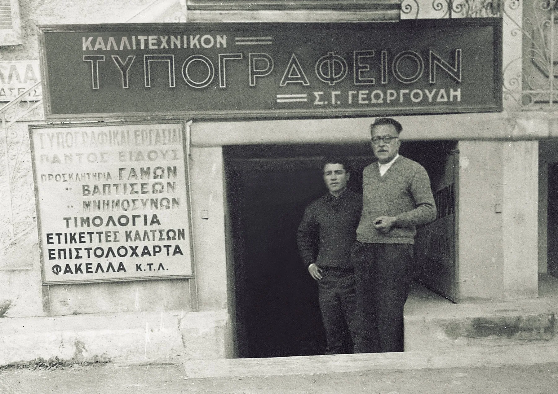 Το τυπογραφείο Σ. Γ. Γεωργούδη: δύο άνδρες στην είσοδο, κάτω από την επιγραφή «Καλλιτεχνικόν Τυπογραφείον»